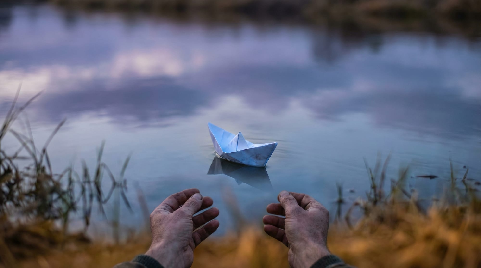 Hands releasing a paper boat onto a still river at dusk